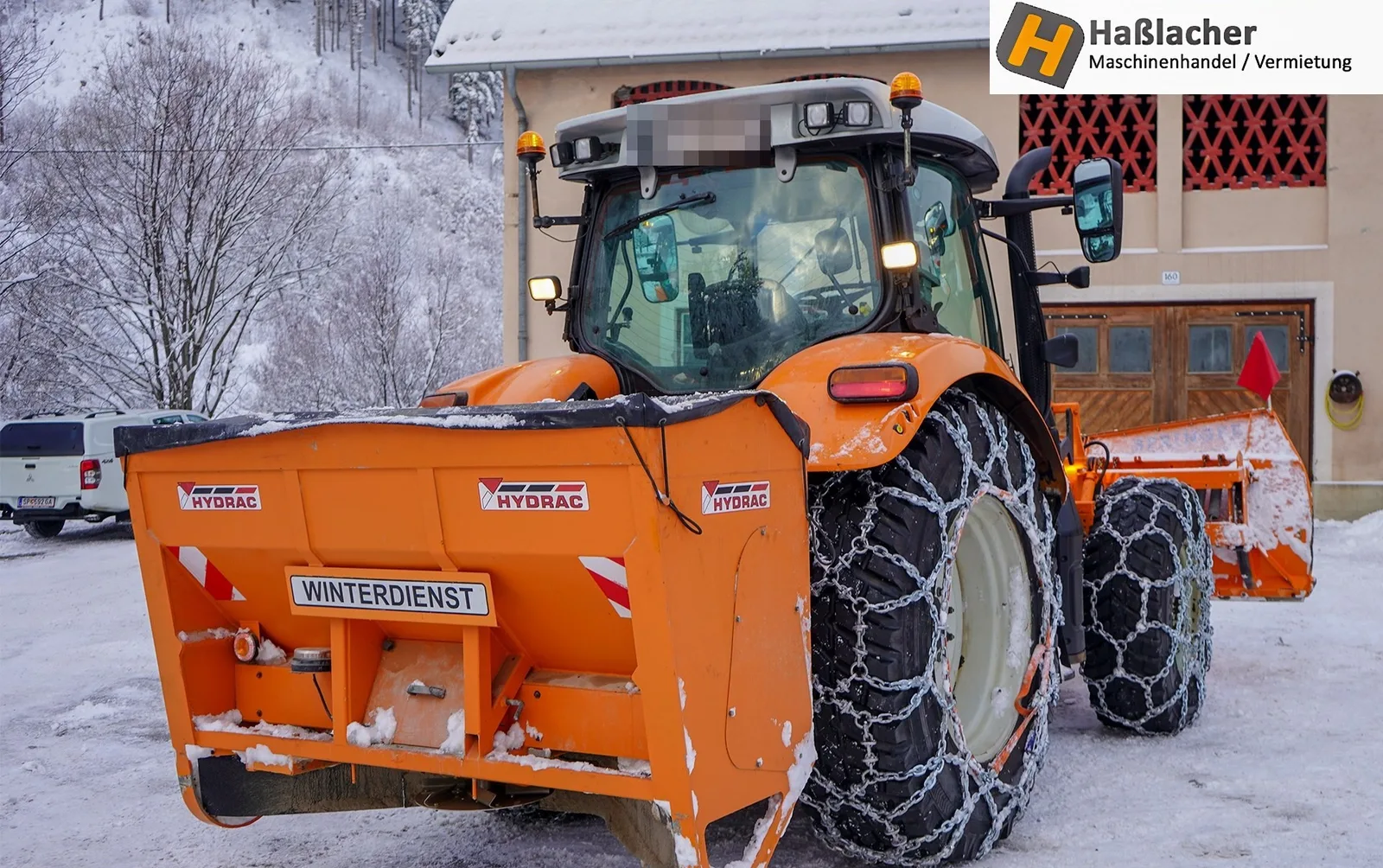 Oranger Traktor mit Winterdienst-Streuer und Schneeketten im verschneiten Hof