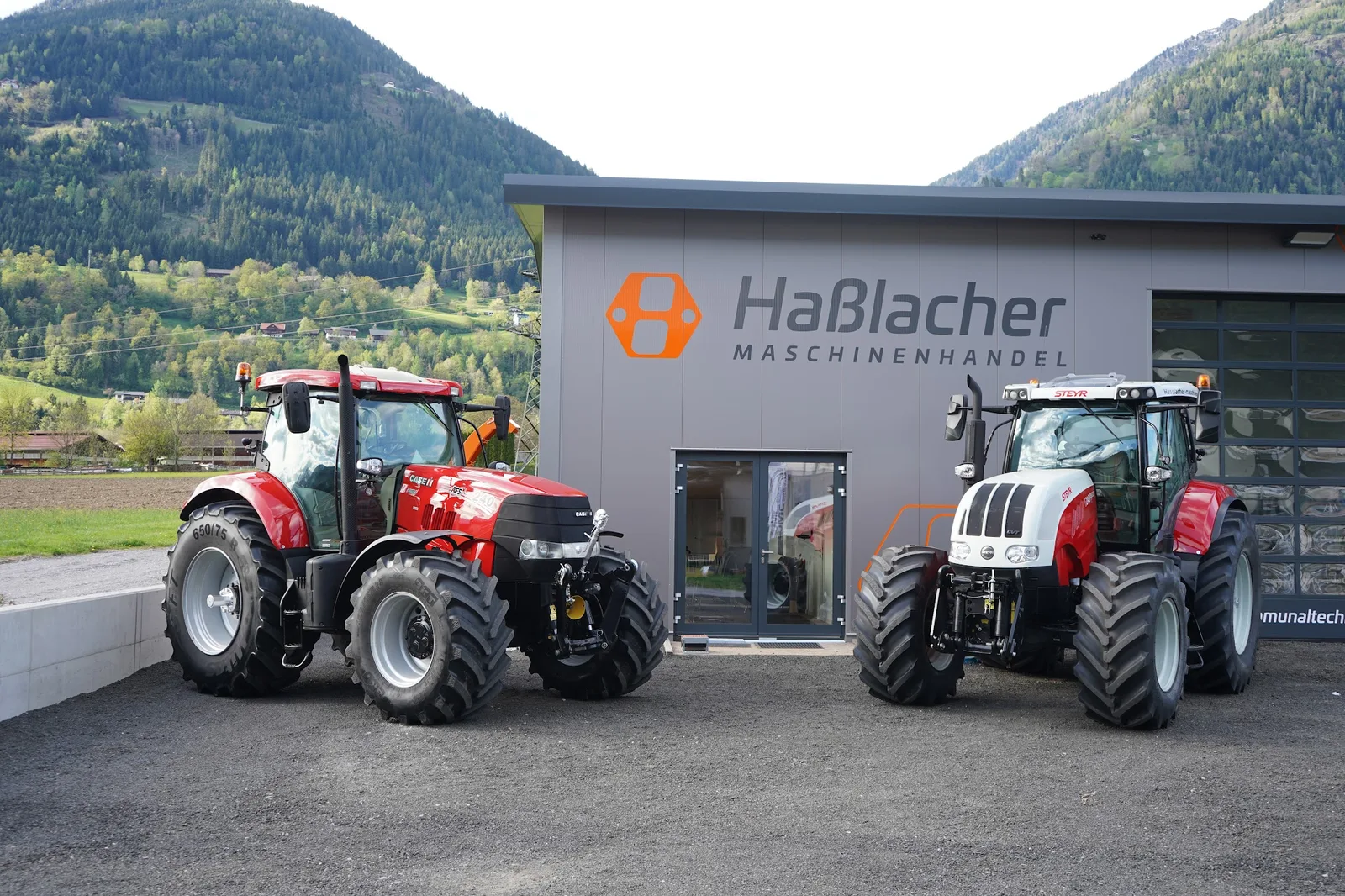 Halle von Haßlacher Maschinenhandel mit Logo, davor ein roter Case IH und ein weiß-roter Steyr Traktor, dahinter bewaldete Berge