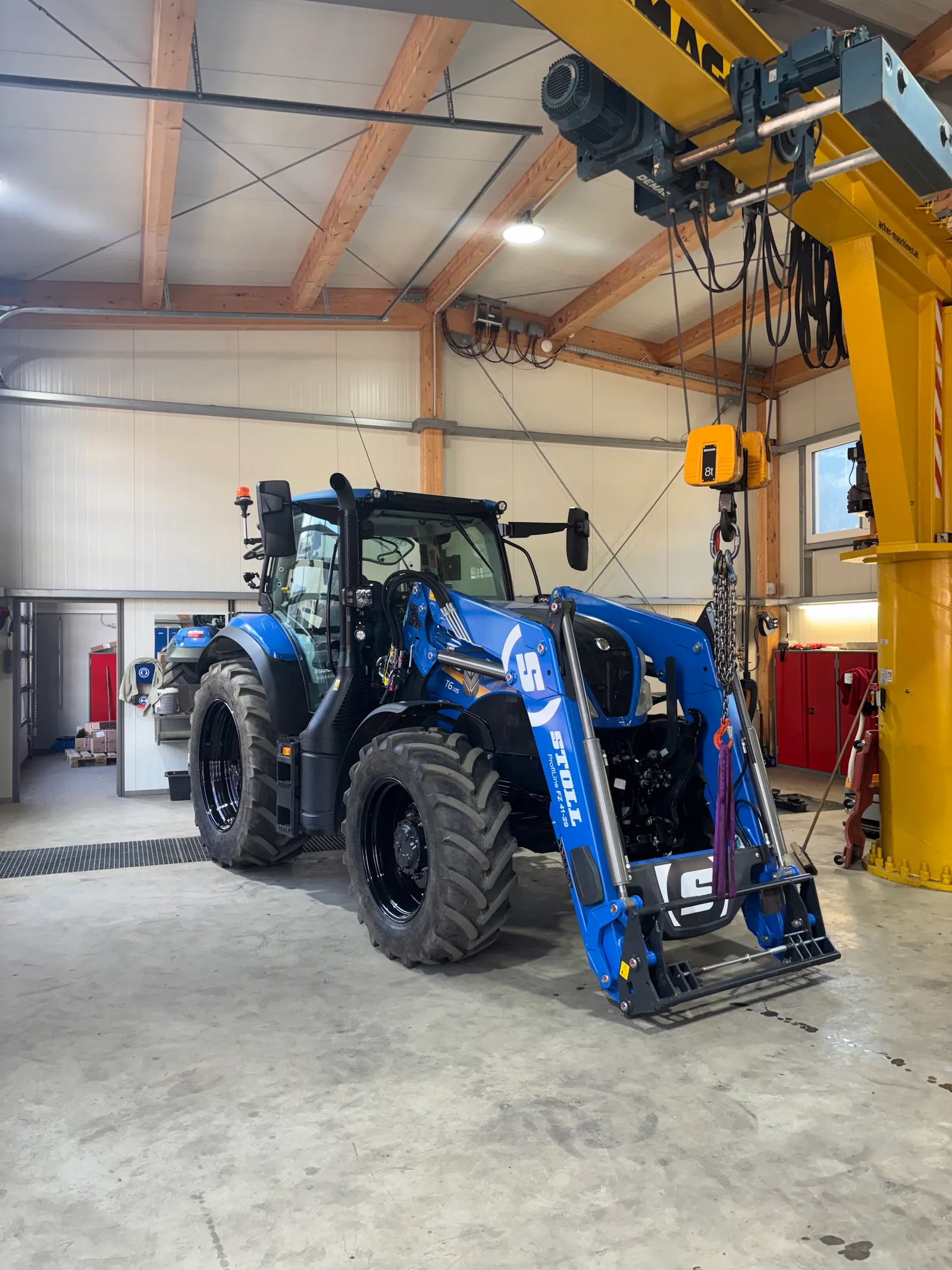 Blauer New Holland Traktor mit Stoll Frontlader in der Werkstatthalle unter dem Hallenkran