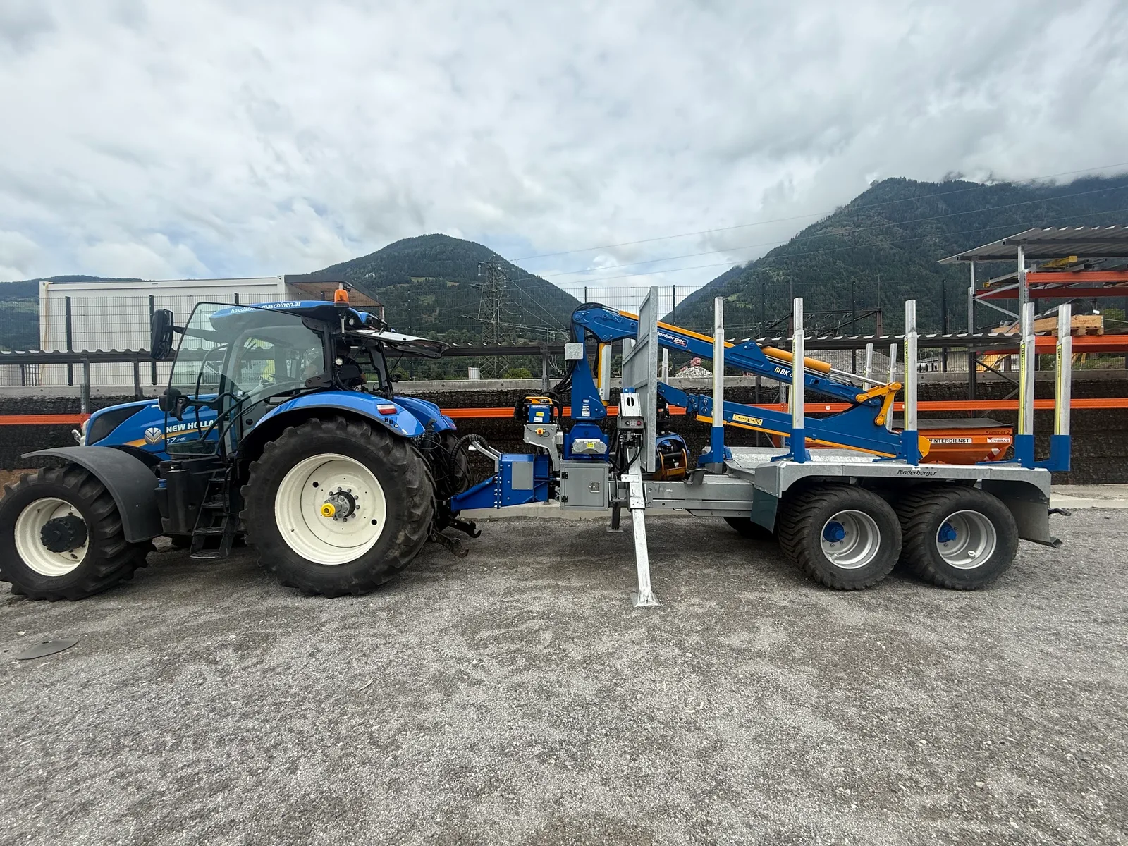 Blauer New Holland Traktor mit angehängtem Forstanhänger und Ladekran am Hof, dahinter Berge
